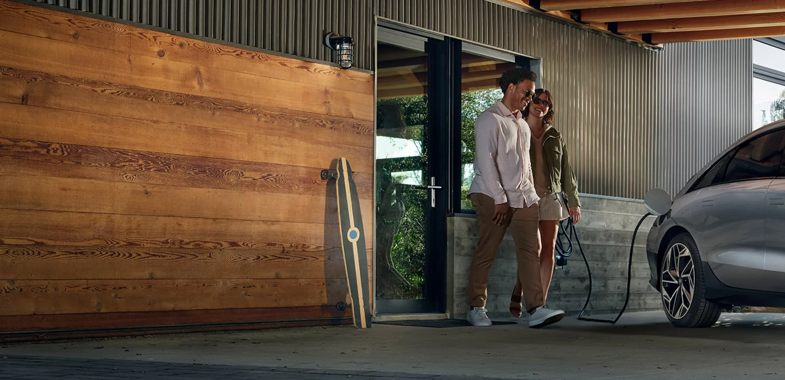 A couple walks past a modern house with a wooden garage door and a silver electric car charging outside.