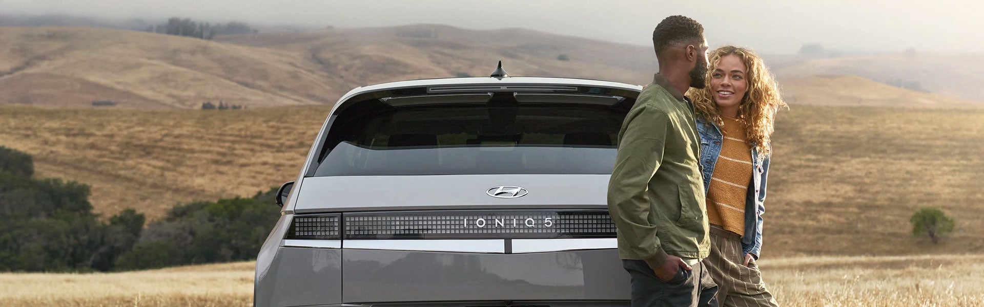 A couple stands next to a gray Hyundai IONIQ 5 SUV, parked on a dirt road with rolling hills in the background.