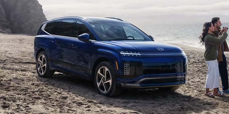 Blue Hyundai IONIQ 9 SUV parked on a sandy beach near the ocean, with two people standing beside the vehicle