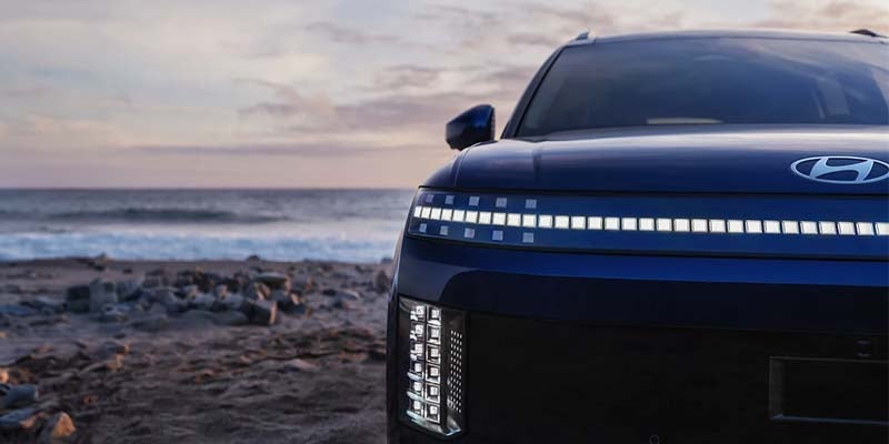 Close-up front view of a blue Hyundai IONIQ 9 SUV on a beach, highlighting the pixel-style LED light bar and distinctive front lighting design