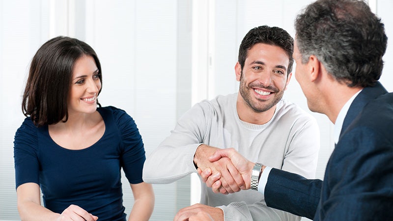 Three people seated at a table during a business meeting, with two individuals shaking hands while discussing a financial
